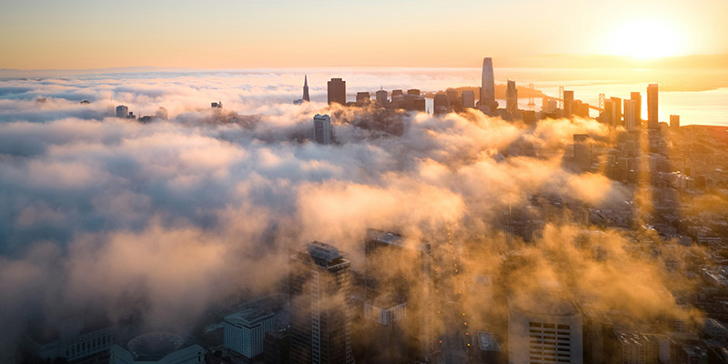 Aerial view of a city skyline at sunset, partially obscured by fog. The sun is setting behind tall buildings, casting warm light over the scene.