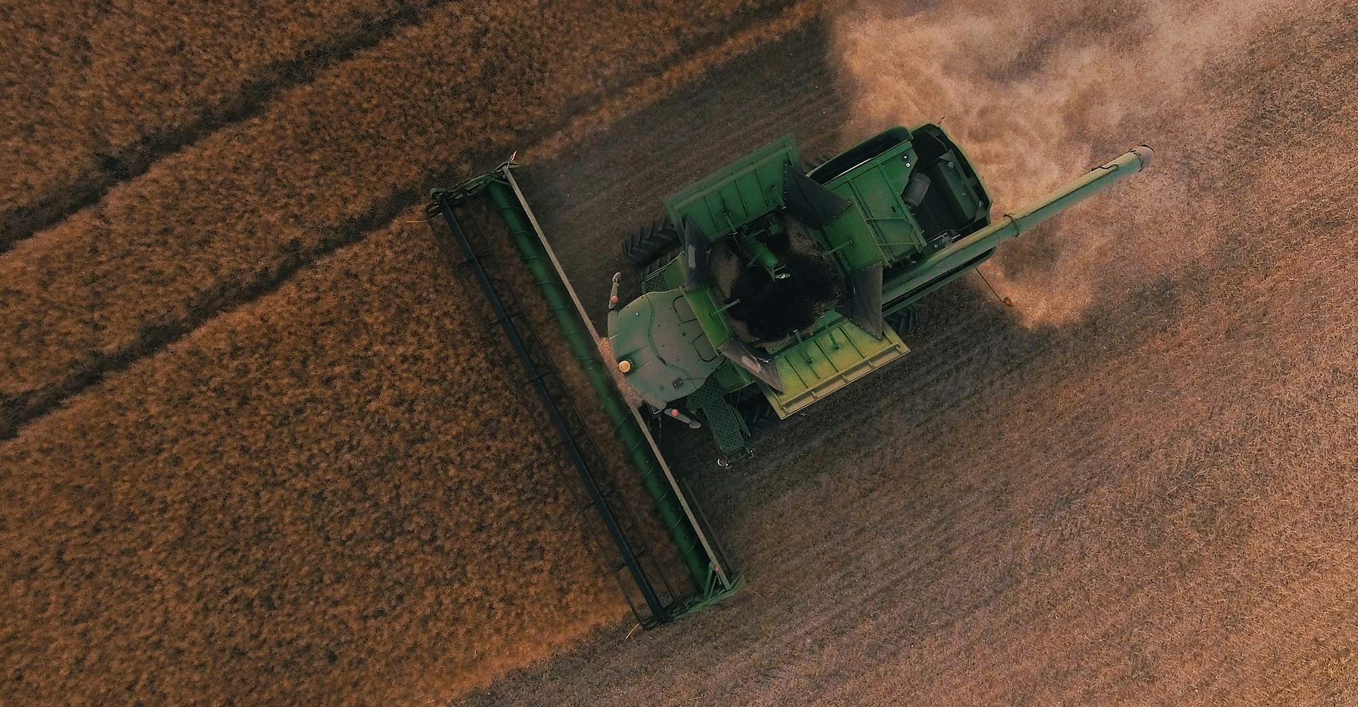 Aerial view of a green combine harvester cutting through a field of crops, producing dust behind it, creating distinct lines in the field.