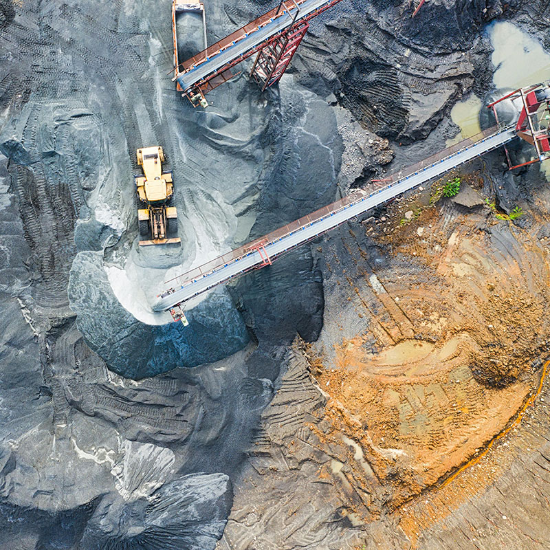 Aerial view of a mining site with heavy machinery and conveyor belts moving materials, displaying a mix of gravel, sand, and water puddles on the ground.