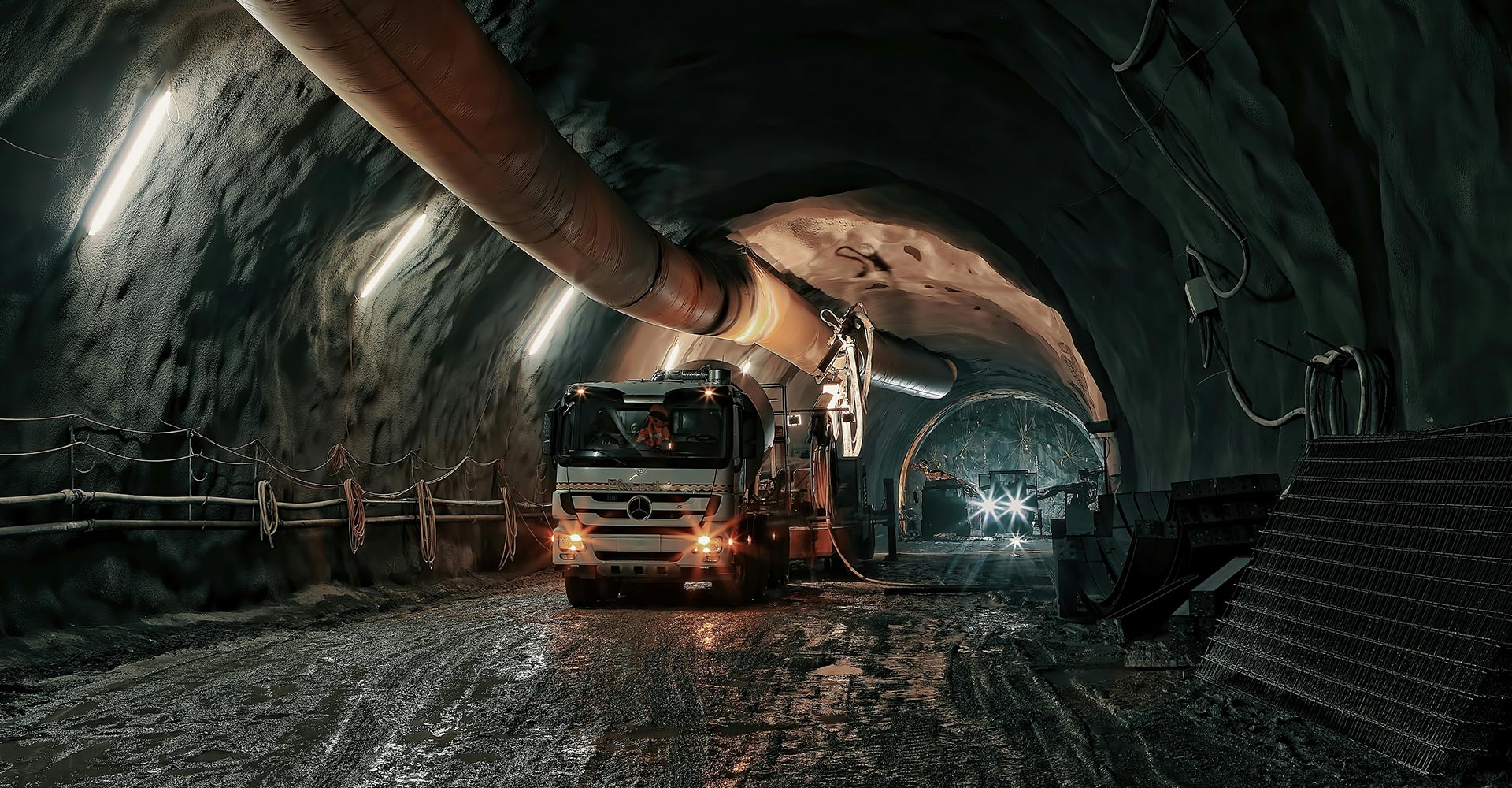 A large tunnel with illuminated light strips, where industrial vehicles operate on the muddy ground. A truck with equipment attached is prominent, displaying headlights and work lights.