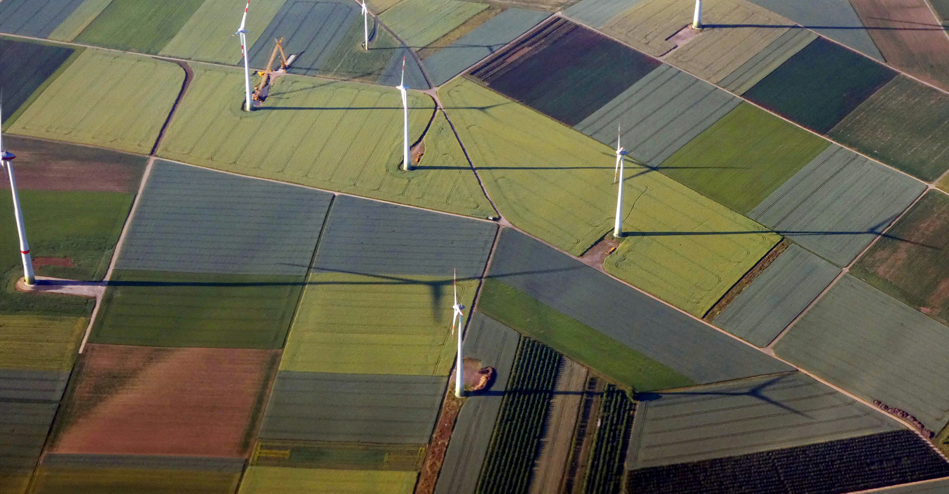 Aerial view of a patchwork of agricultural fields with several wind turbines casting long shadows across the landscape.